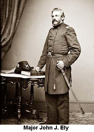 Black and white photograph of Union Major John J. Ely, standing, with his right hand on the hilt of a dress saber and his left hand resting on a book which is placed on an ornate carved wooden table; his dress uniform hat is leaning against the book